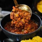 A helping of turkey chili being ladled into a dark blue bowl next to a piece of cornbread.