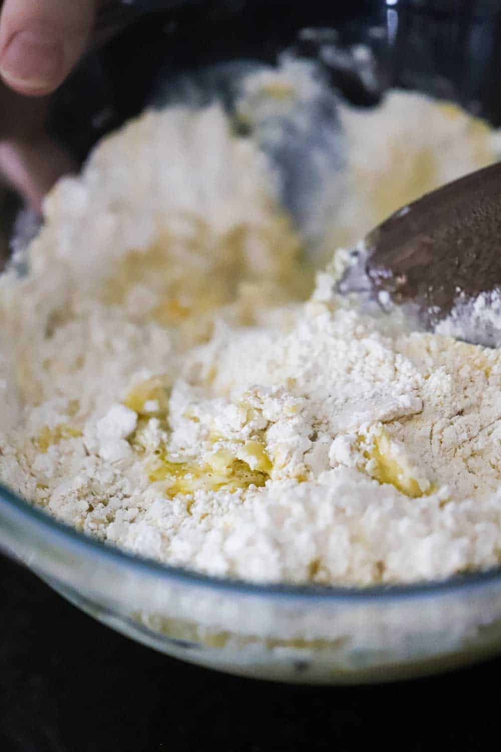 A glass bowl filled with flour, water, yeast, and cornmeal all being mixed together by a wooden spoon. 