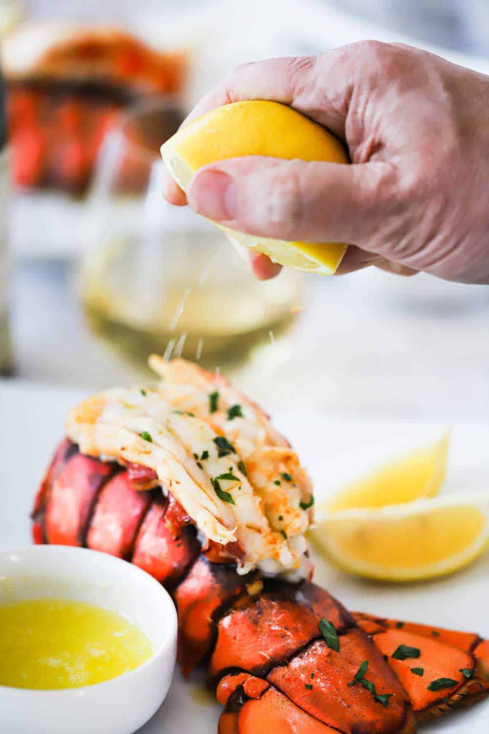 A person squeezing a half of a fresh lemon over a cooked lobster tail sitting next to a small bowl of drawn butter. 