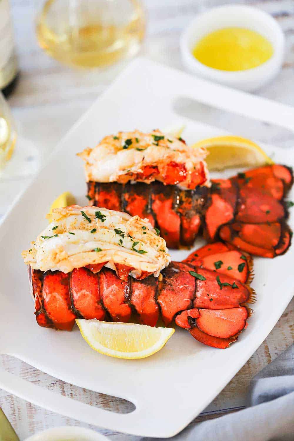 Two fully cooked lobster tails with meat on top of the lobster tail, all sitting on a platter next to lemon wedges and melted butter. 