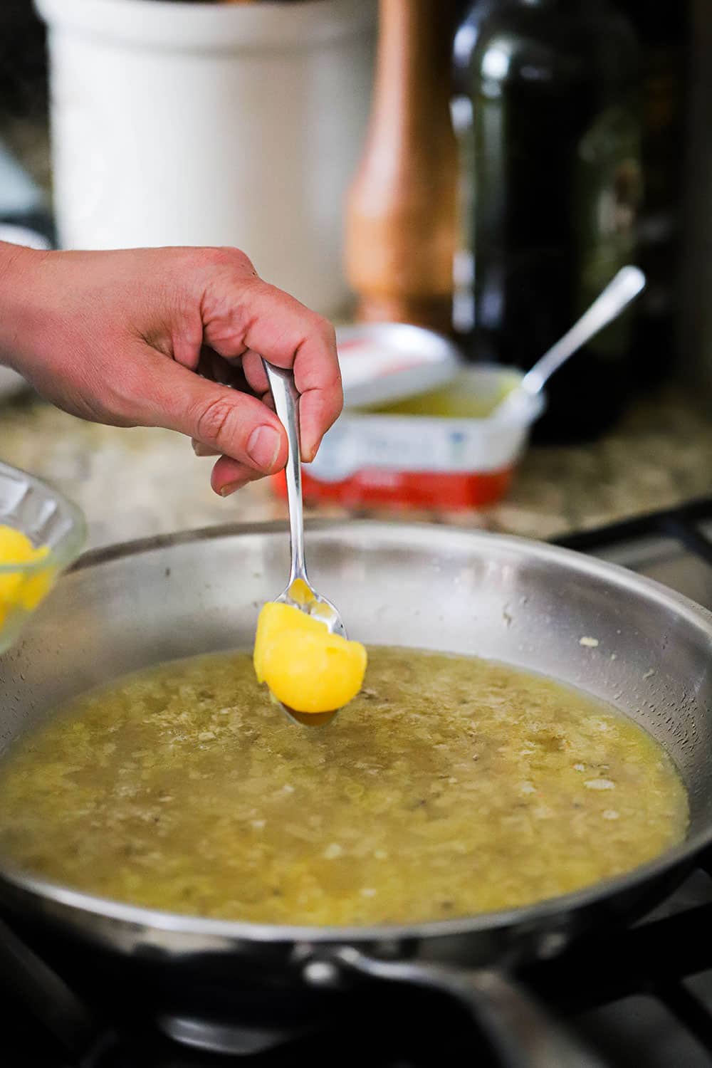 A person holding a spoon filled with clarified butter over skillet filled with a white wine sauce.