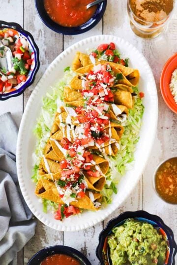 An oval white platter with a layer of shredded lettuce on the bottom topped with a row of chicken flautas surrounded by bowls of salsa, guacamole, and pico de gallo.