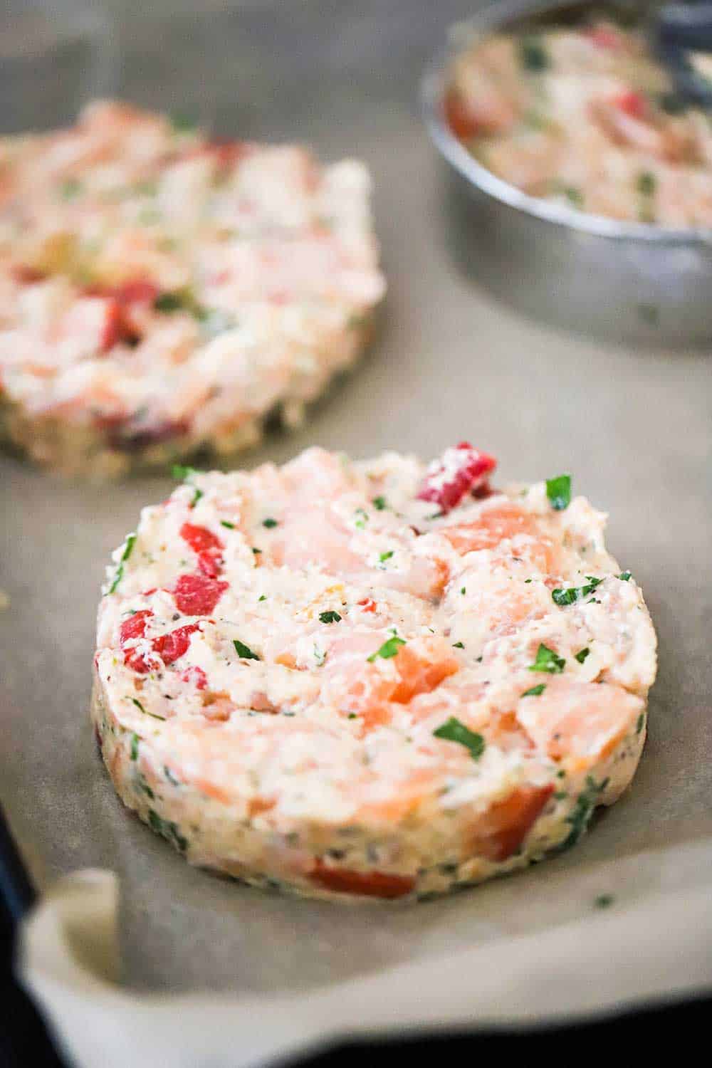 An uncooked salmon burger patty sitting next to other patties on parchment paper. 