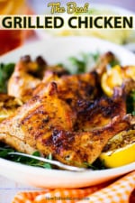 A grilled chicken leg quarter sitting on a platter next to grilled lemons and fresh herbs.
