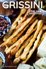 A pile of baked grissini, also known as Italian breadsticks, on a board next to a block of cheese that has been sliced and a bowl of spiced nuts.
