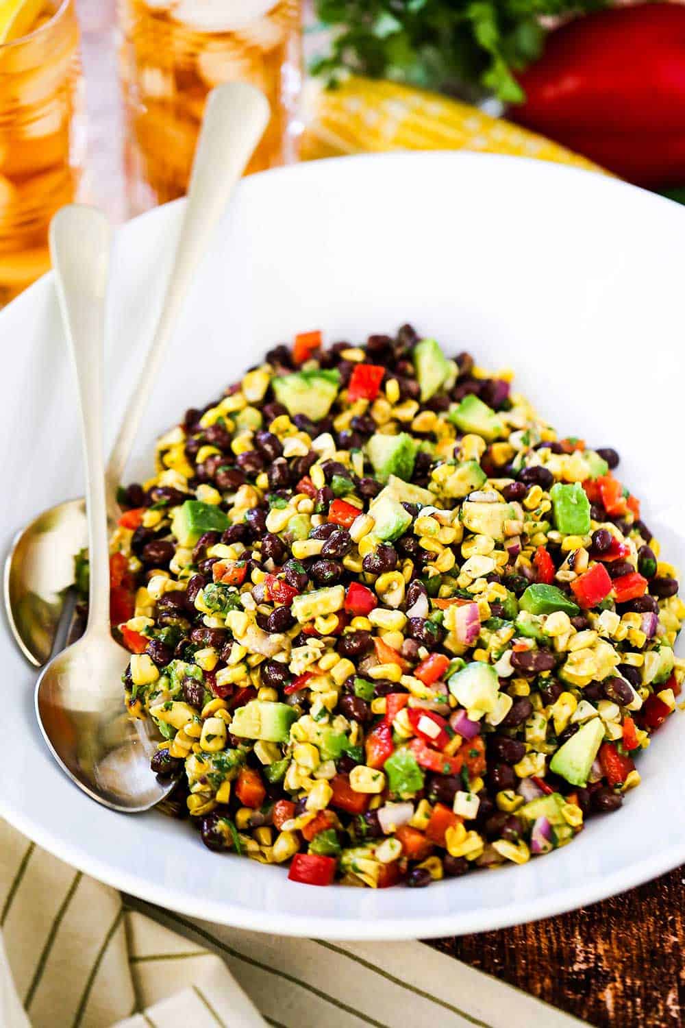 A large white serving bowl filled with black bean and corn salad with two gold serving spoons tucked in beside the salad.