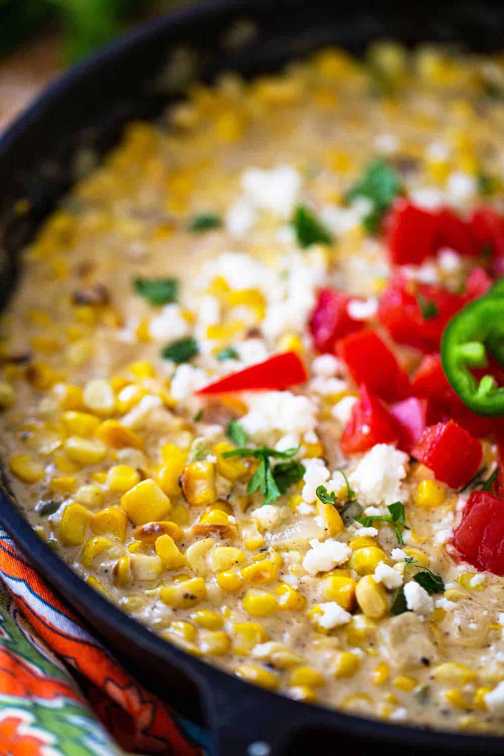 A cast-iron skillet filled with Mexican cream corn topped with crumbled queso fresco, chopped tomatoes, and chopped cilantro.