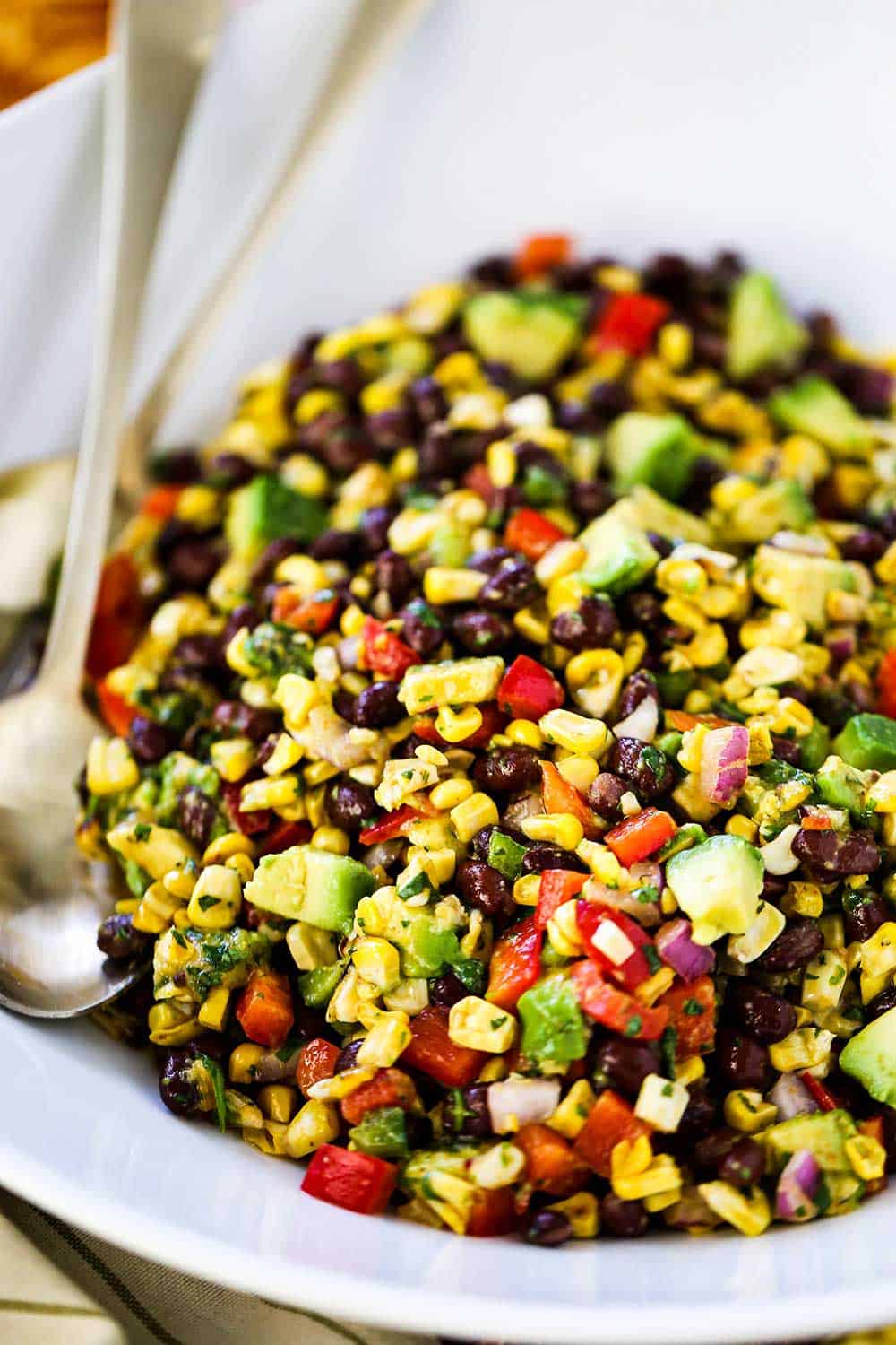 A large white serving bowl filled with black bean and corn salad with two gold serving spoons tucked in beside the salad.