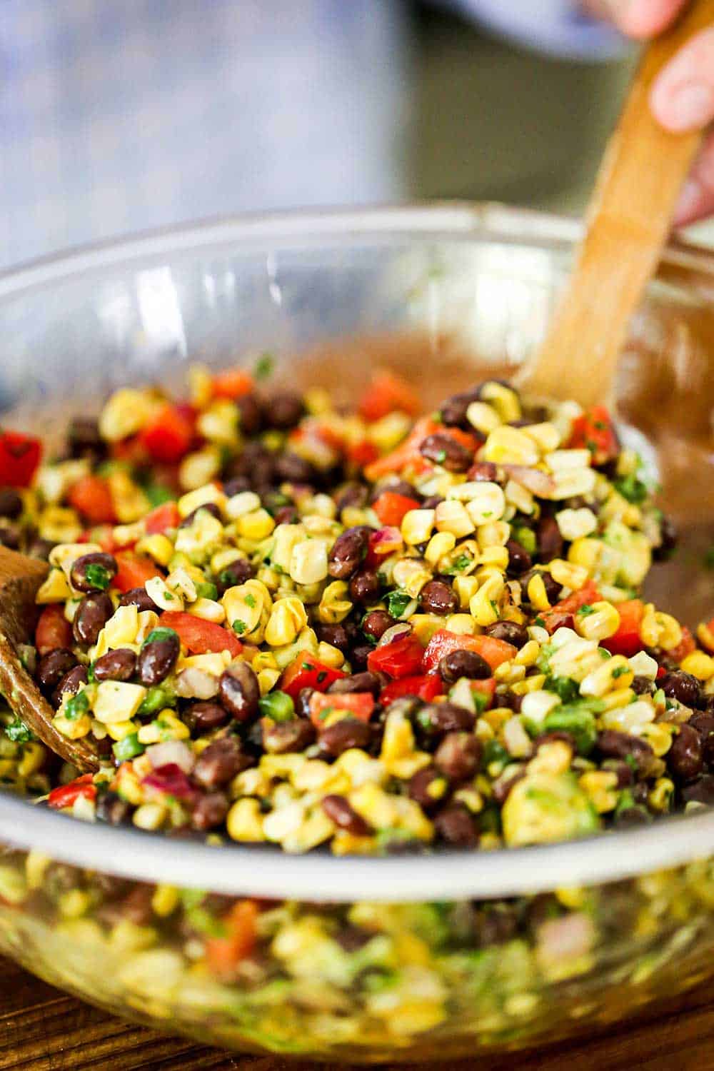 A glass bowl filled with fresh corn, black beans, red bell peppers, and avocado pieces all being stirred by a wooden spoon.