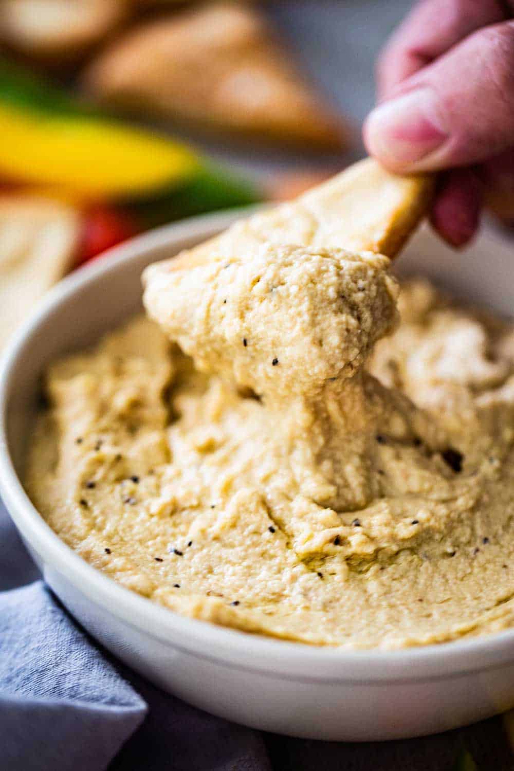 A person lifting a baked pita chip out of a bowl of homemade hummus. 