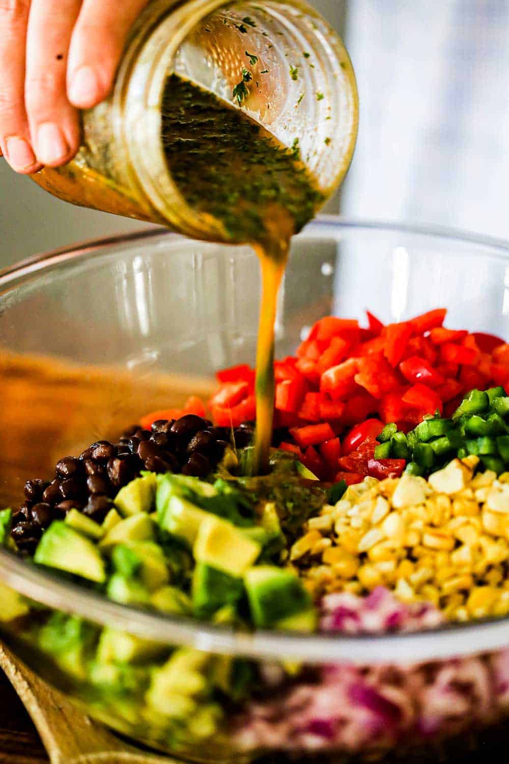 A person pouring a cilantro lime dressing from a glass jar into a glass bowl filled with black beans and cut vegetables.