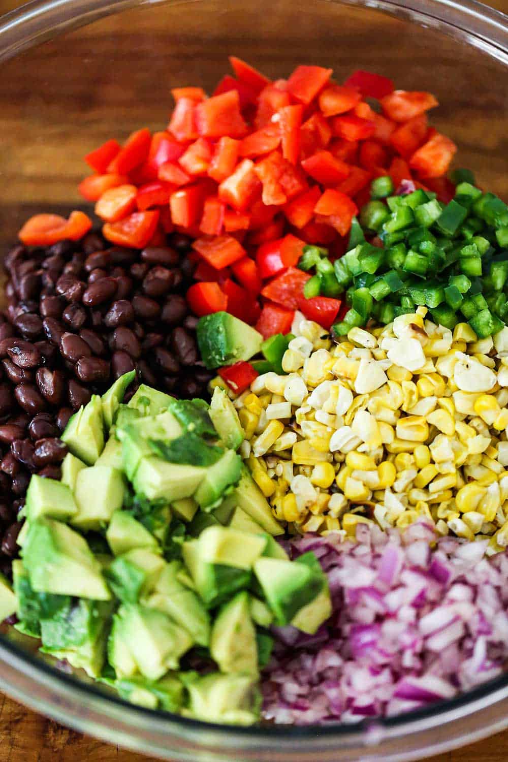 A glass bowl filled with a portion each of chopped avocado, red onion, corn kernels, jalapeno, red bell peppers, and black beans.
