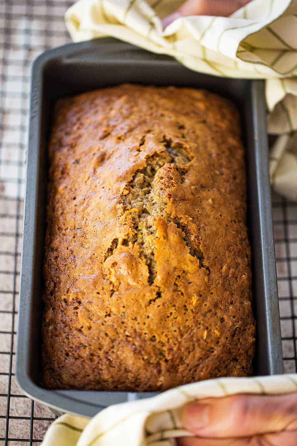 A pair of hands using two kitchen linens to hold a pan filled with freshly baked zucchini bread. 