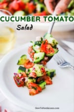 A person holding a large serving spoon filled with cucumber tomato salad over a white dinner plate with a helping of the salad on it.