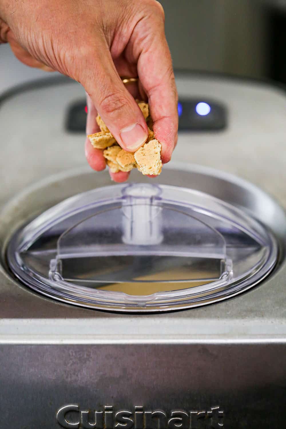 A person using their fingers to drop chopped pieces of graham crackers into the metal pot of an ice cream maker.