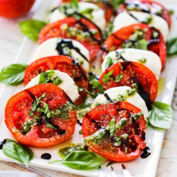 A white rectangular platter holding layers of slice tomatoes and mozzarella to form a caprese salad being topped with squirts of pesto sauce from a squeeze bottle.