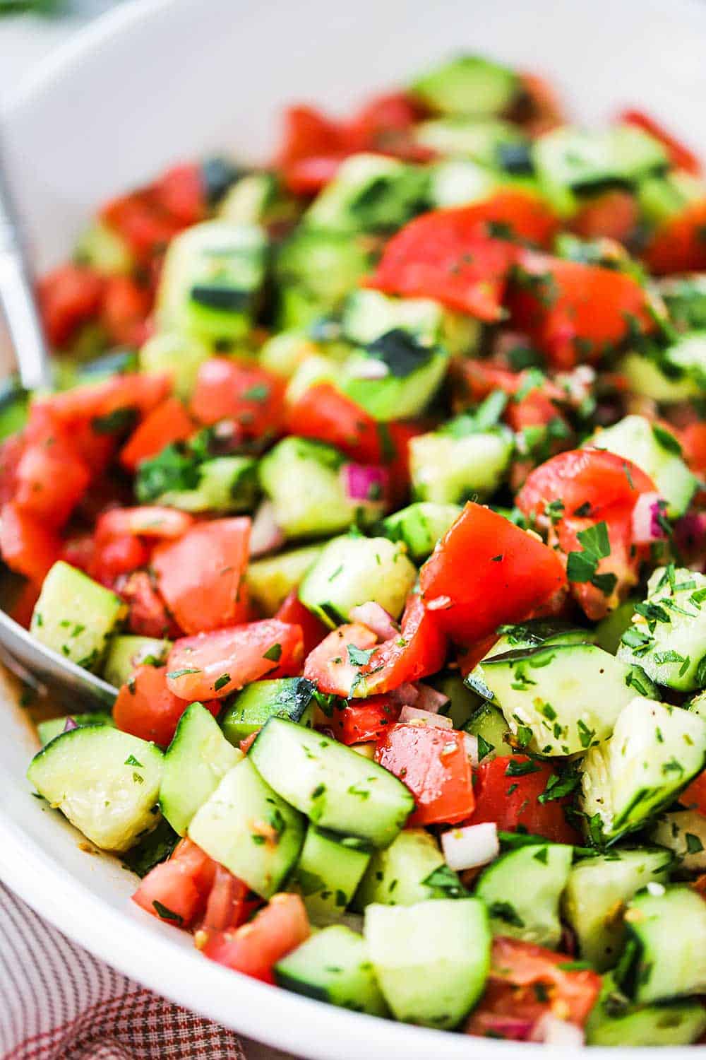 A cucumber tomato salad in a large oval white serving dish with a silver serving spoon stuck in the side. 