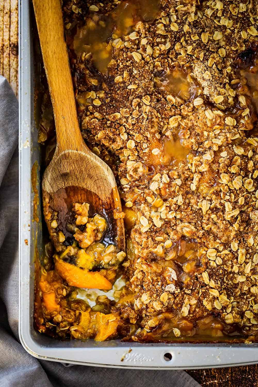 A silver baking pan filled with fully baked peach crisp with a wooden spoon inserted in the side.