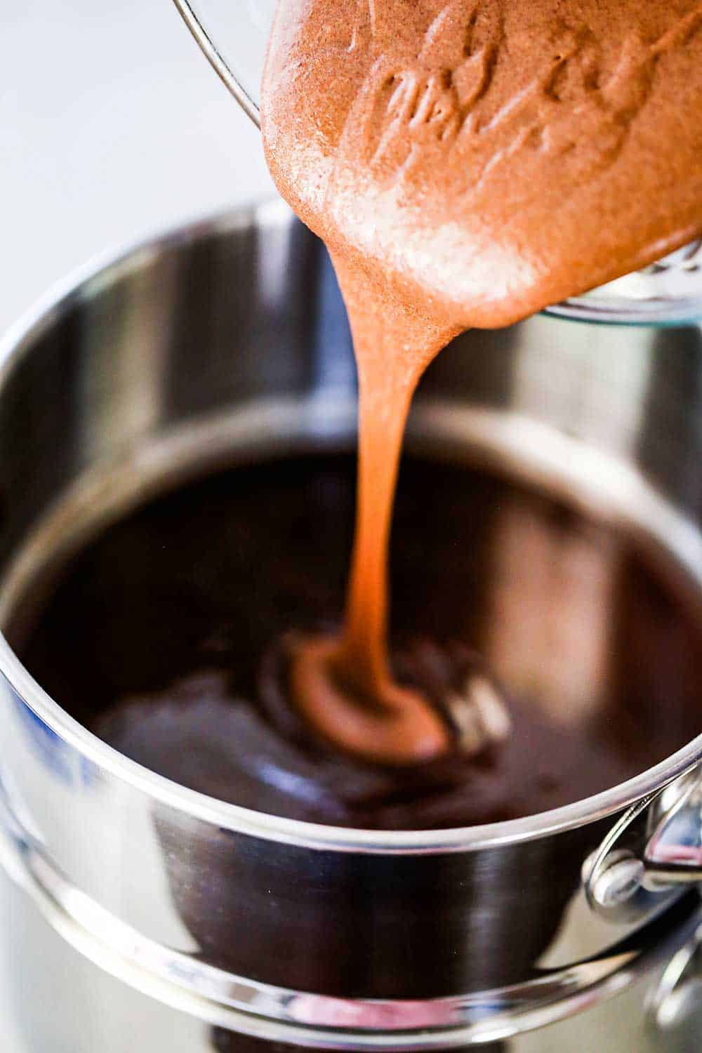 A creamy chocolate mixture being transferred from a glass bowl into a double boiler filled with melted butter and chocolate.
