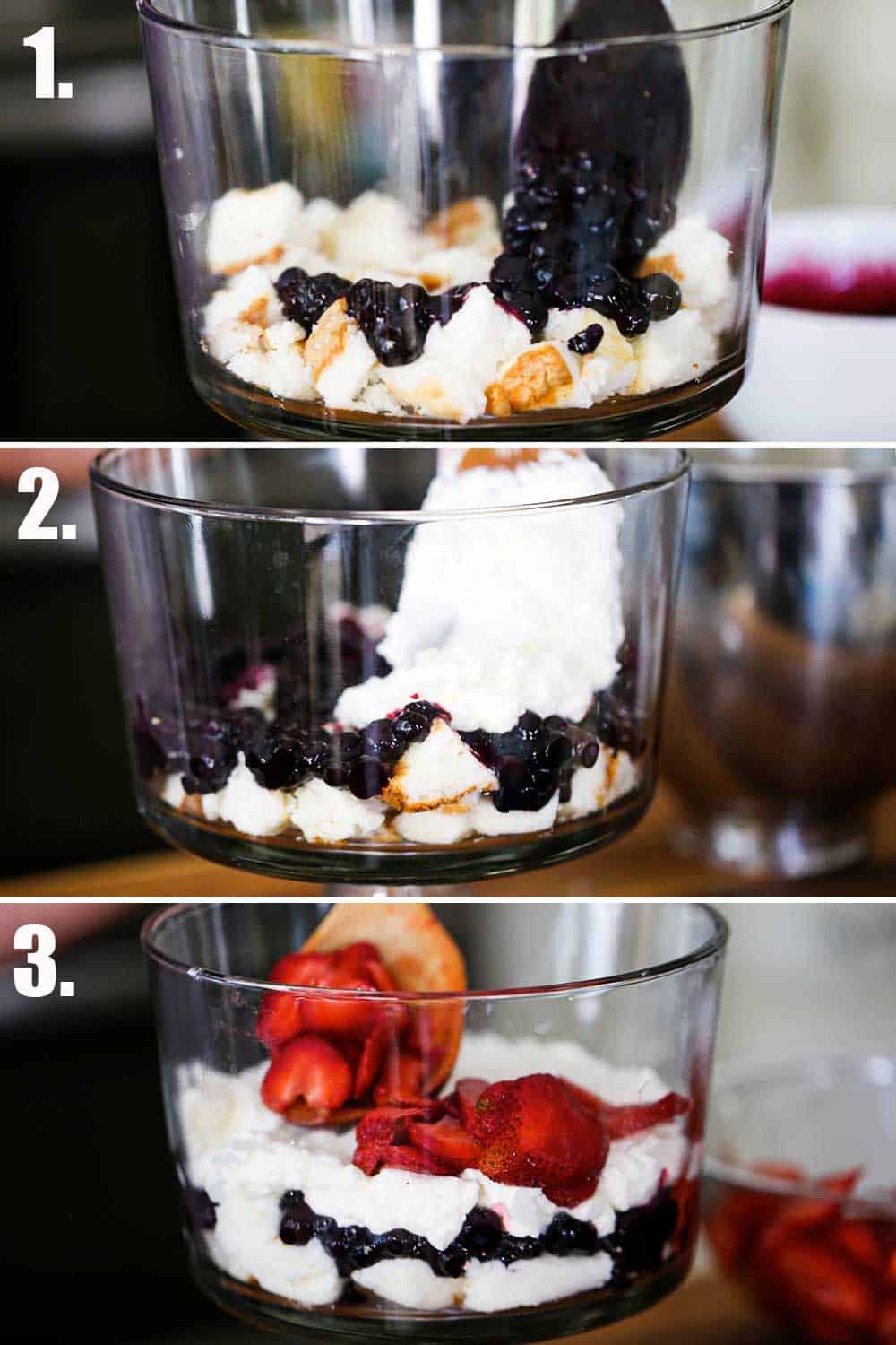 A spoon layering a blueberry sauce over pieces of angel food cake in a trifle dish, and then cream being spooned over the blueberries, and then strawberries being added.