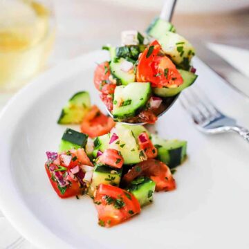 A person holding a large serving spoon filled with cucumber tomato salad over a white dinner plate with a helping of the salad on it.