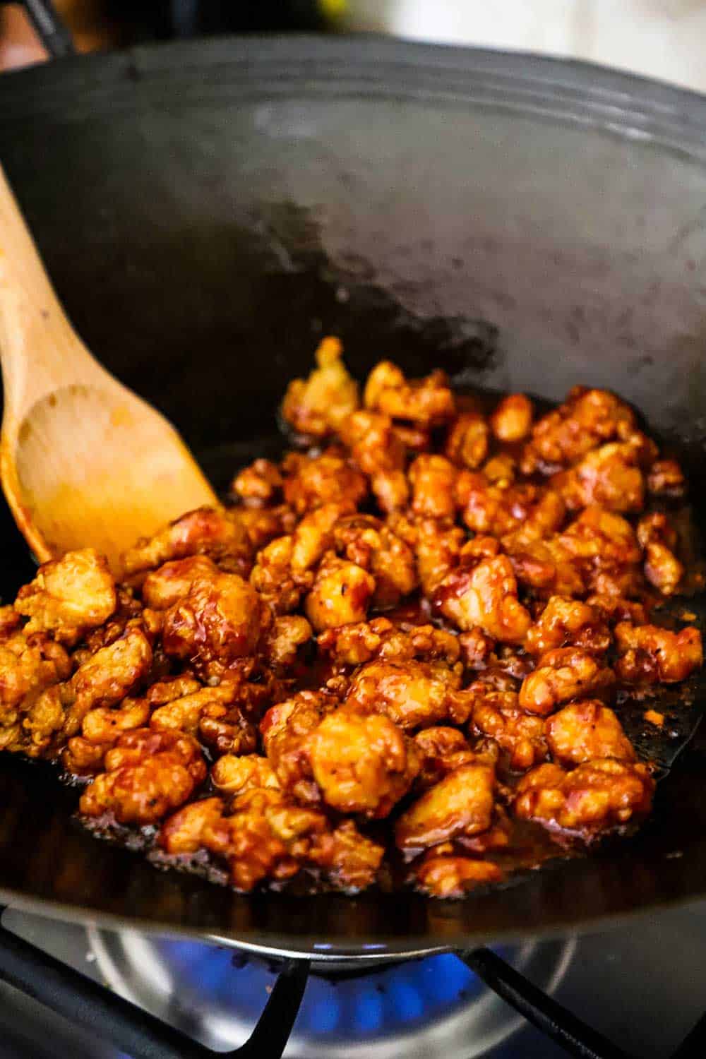 A wok on a gas-burning stove filled with sesame chicken and wooden spoon.