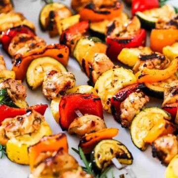 A row of 5 chicken kabobs on a white oval platter garnish with fresh rosemary sprigs and a bowl of white sauce.