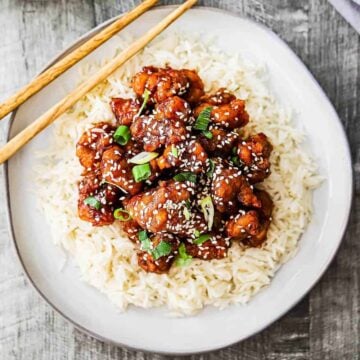 A dinner plate filled with a bed of white rice with sesame chicken on top of it and a pair of chop sticks nearby.