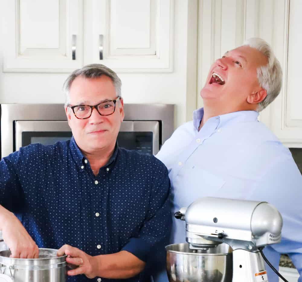 A married couple in the kitchen with one man looking straight-ahead near a container of flour and his husband standing next to him laughing.