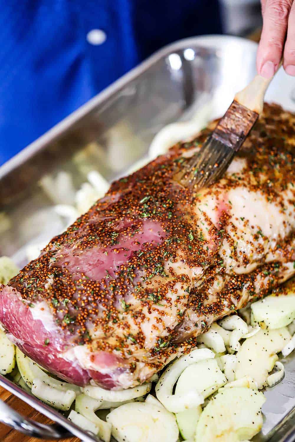 A person using a cooking brush to apply a mustard maple sauce over a large pork loin roast in a roasting pan.