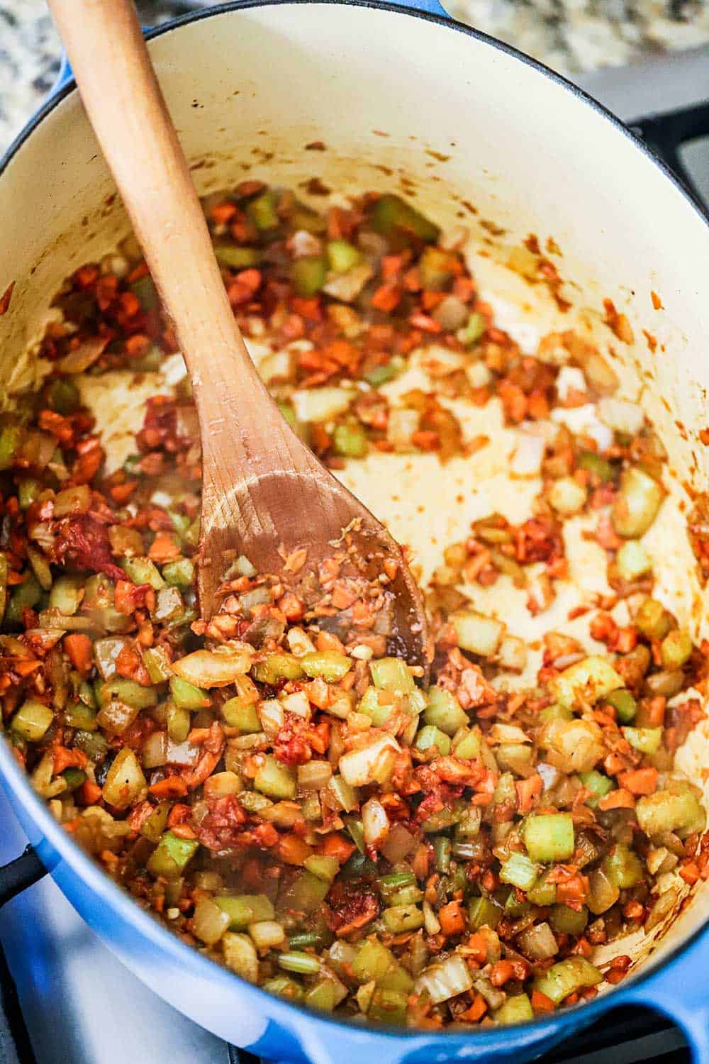 A large blue Dutch oven filled with a mirepoix of chopped onion, carrots, and celery with a wooden spoon in the middle of it all.