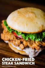 A chicken fried steak sandwich with cream gravy oozing from the bottom bun sitting on a cutting board.