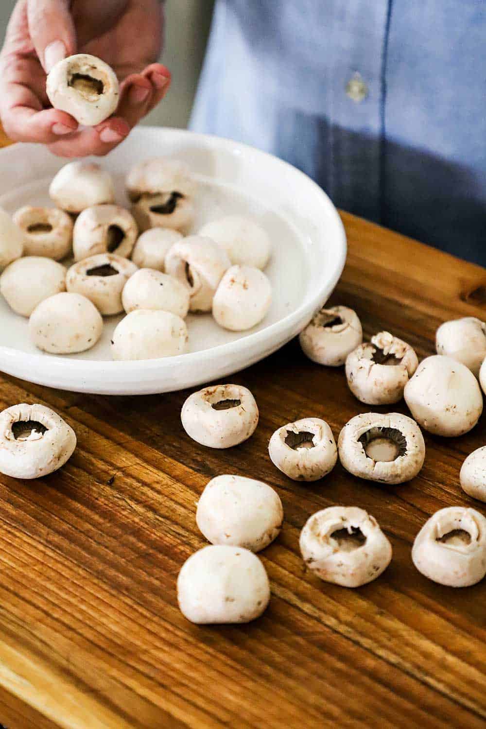 A person holding a white button mushroom with the stem missing over a bowl and cutting board filled with other stemless mushrooms.