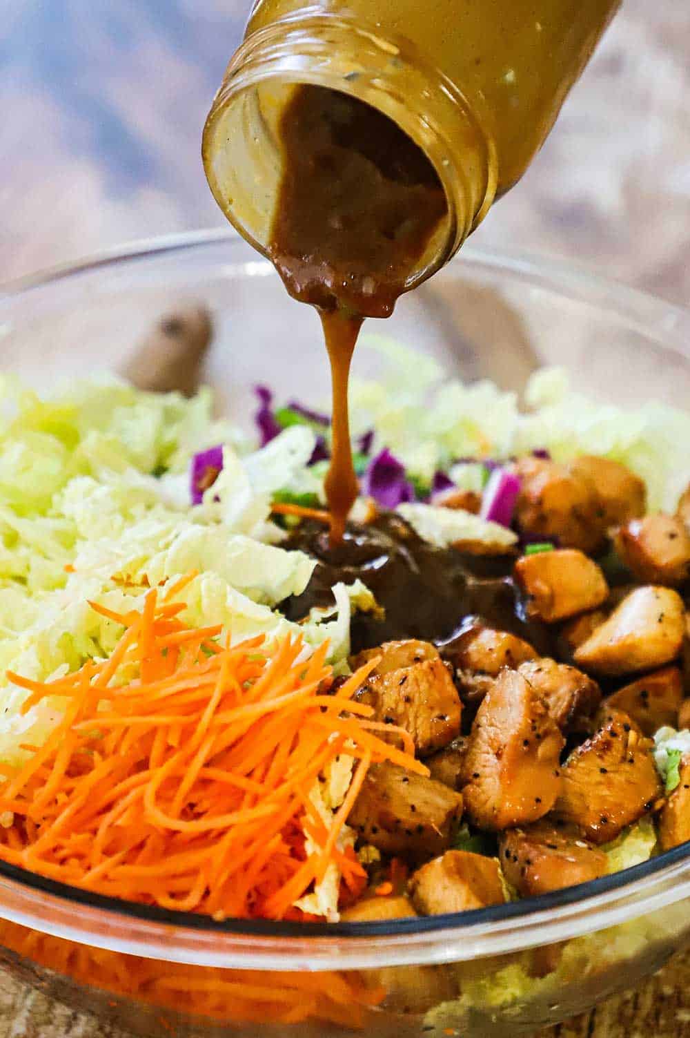 Peanut dressing being poured from a glass jar over a platter filled with chopped vegetables and cooked chicken pieces. 