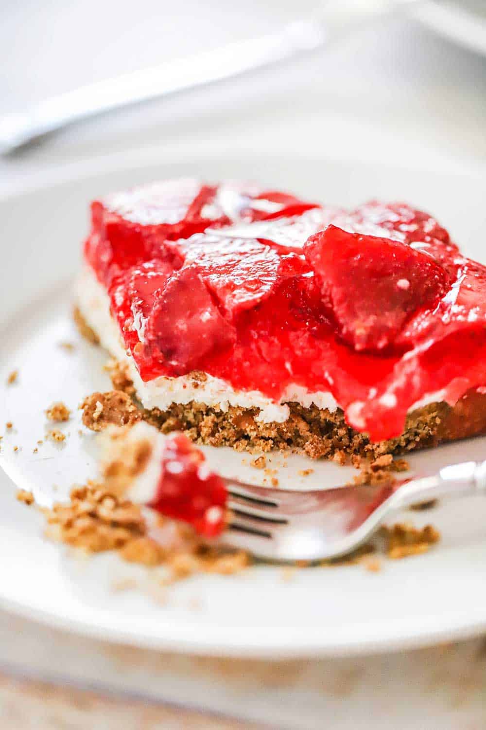 A slice of strawberry pretzel salad with a bite taken out of it sitting on a white plate with a fork next to it. 