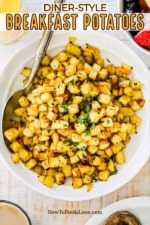 A white serving bowl filled with breakfast potatoes garnished with chopped parsley and with a large spoon stuck in the side.