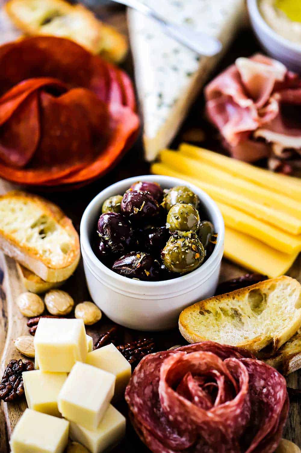 A charcuterie board with meats and cheeses and a white bowl in the middle filled wth marinated olives. 