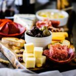 A charcuterie board filled with cubed cheeses, Italian meats in the shape of a flower, and a bowl of hummus.