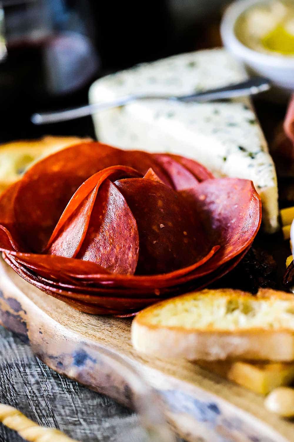 A pile of large pepperoni that has been formed into a flower shape sitting next to a wedge of brie. 