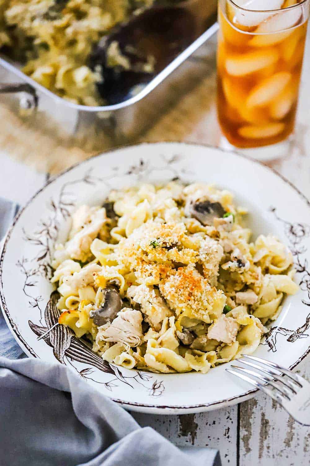 An antique serving bowl filled with a large helping of turkey tetrazzini sitting next to a glass of iced tea. 