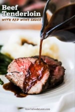 A glass gravy boat pouring red wine sauce over two beef tenderloin slices on a plate along with a pile of mashed potatoes and cooked green beans.
