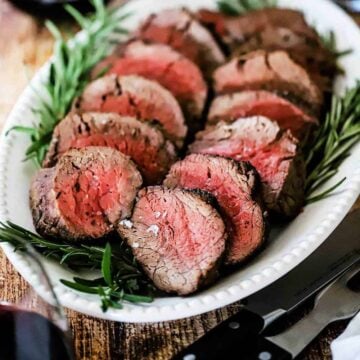 An oval white platter filled with slices of beef tenderloin with sprigs of rosemary along the edge and a glass or red wine sitting in the rear.