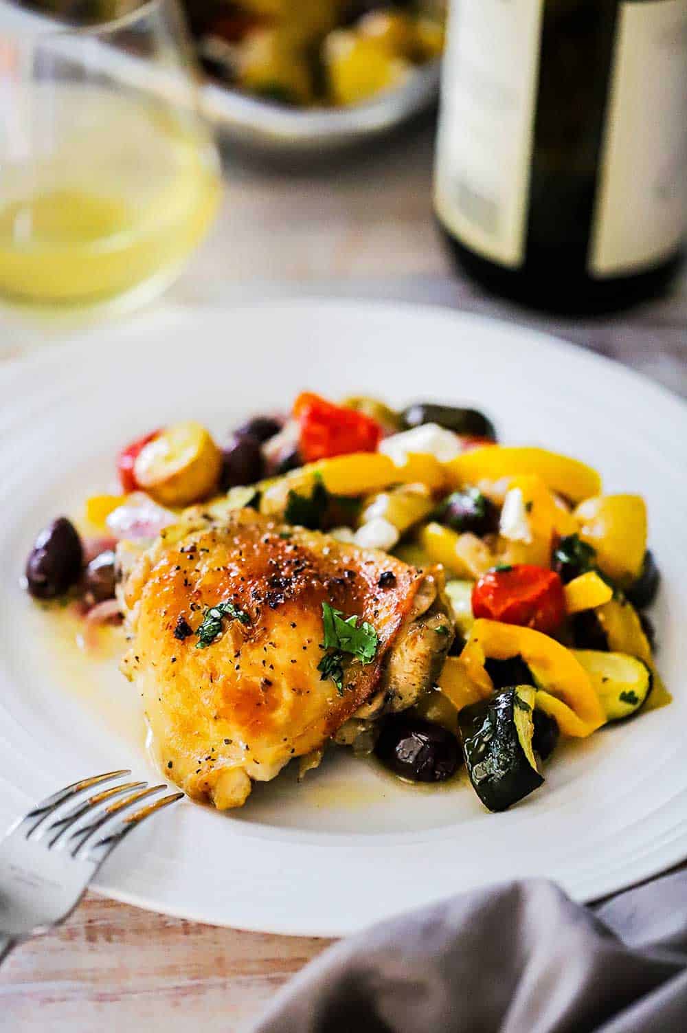 A white dinner plate filled with Sheet Pan Greek Chicken next to a glass of white wine. 