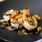 A small dark grey appetizer plate filled with 4 cooked shrimps topped with fried garlic slivers and a white wine and parsley sauce.