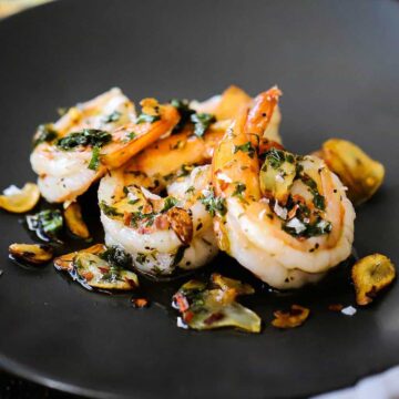 A small dark grey appetizer plate filled with 4 cooked shrimps topped with fried garlic slivers and a white wine and parsley sauce.