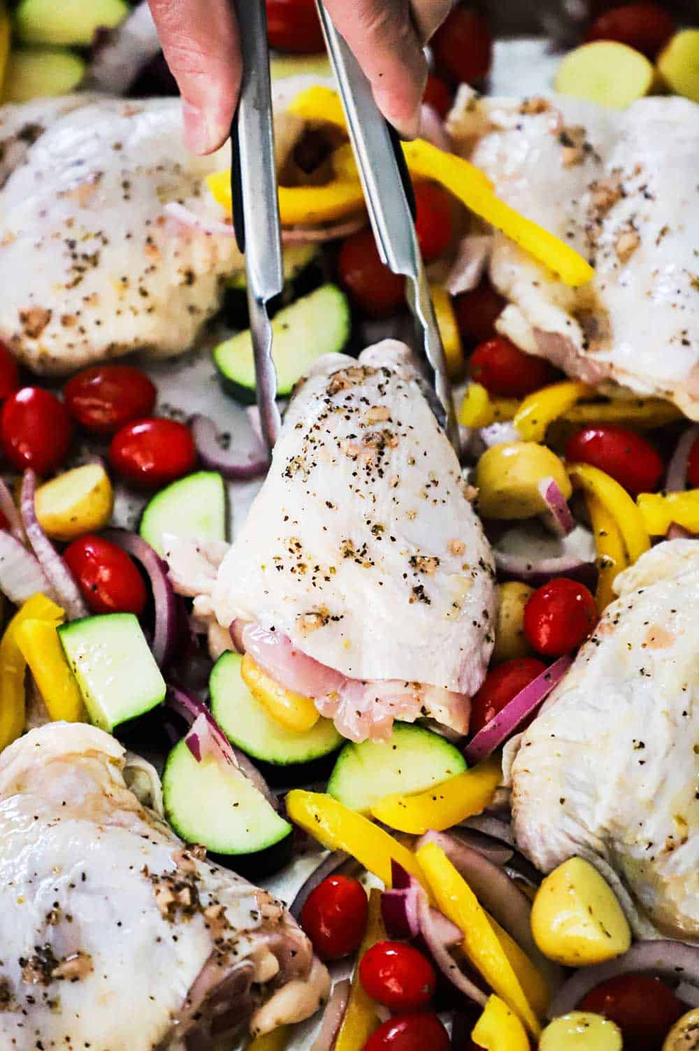 A pair of tongs being used to place an uncooked chicken thigh on a sheet pan with cut vegetables. 