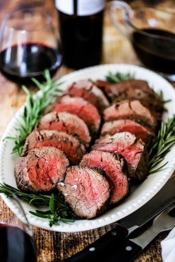 An oval white platter filled with slices of beef tenderloin with sprigs of rosemary along the edge and a glass or red wine sitting in the rear.