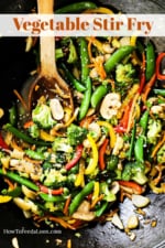 A black metal wok filled with vegetable stir-fry with a wooden spoon stuck in the side.