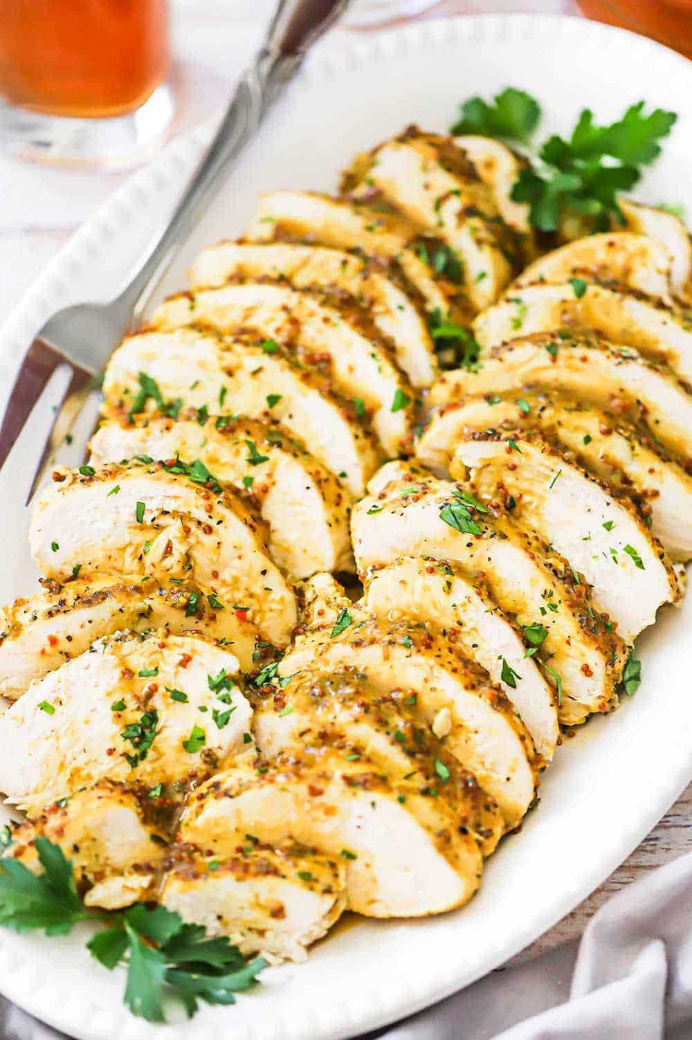 A white oval platter filled with layers of sliced baked honey mustard chicken garnished with sprigs of fresh parsley.