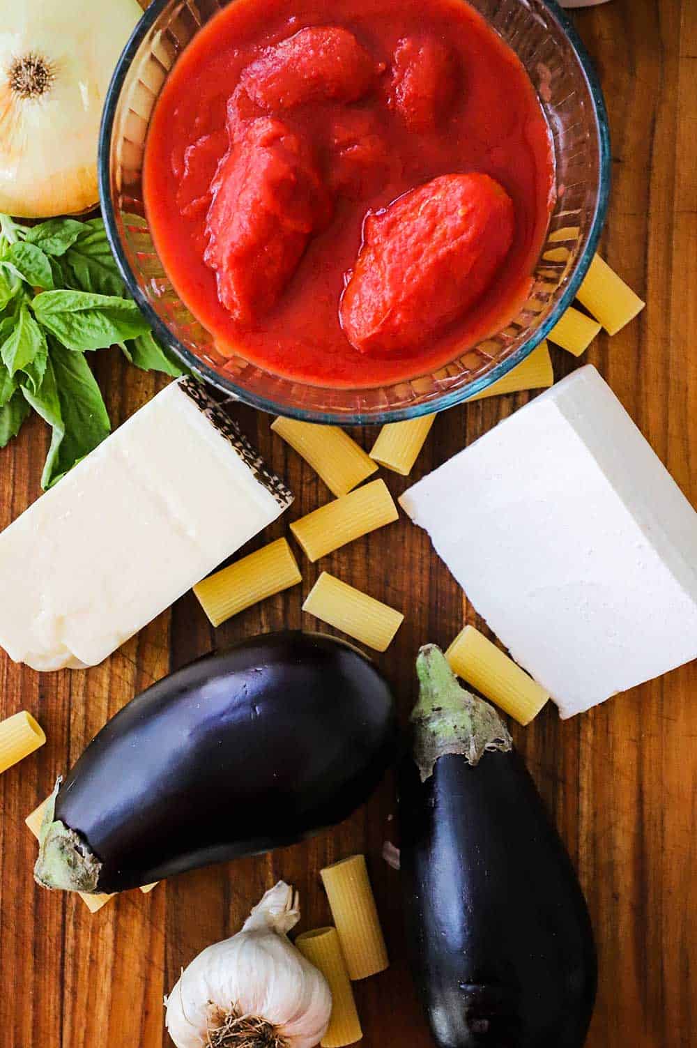 A wooden cutting board with an array of ingredients on it including a bowl of tomatoes, a whole onion, two eggplants, dried past and two pieces of white cheese.